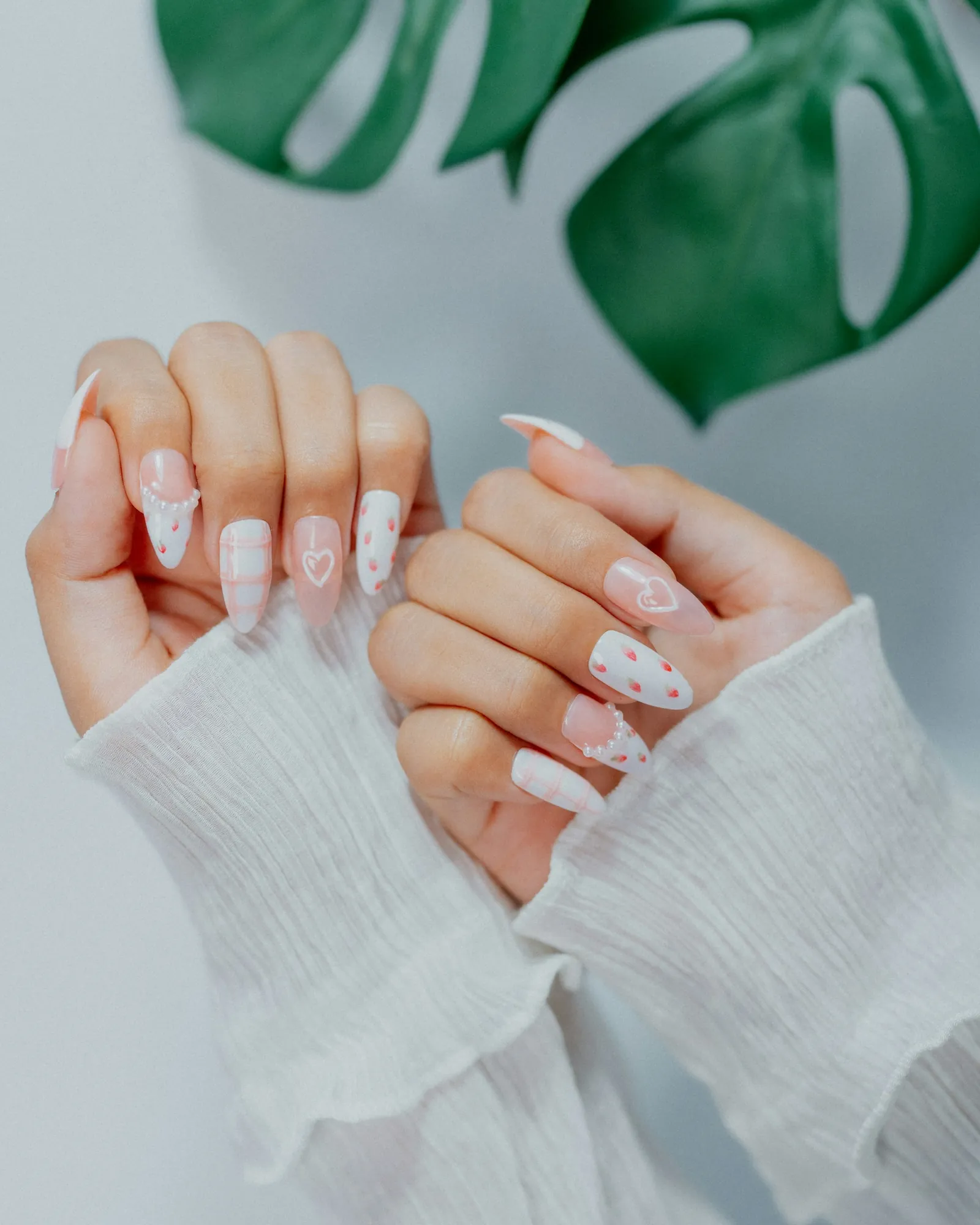 Manos con uñas almendra decoradas con corazones y lunares rosas sobre un jersey blanco, junto a una hoja de monstera